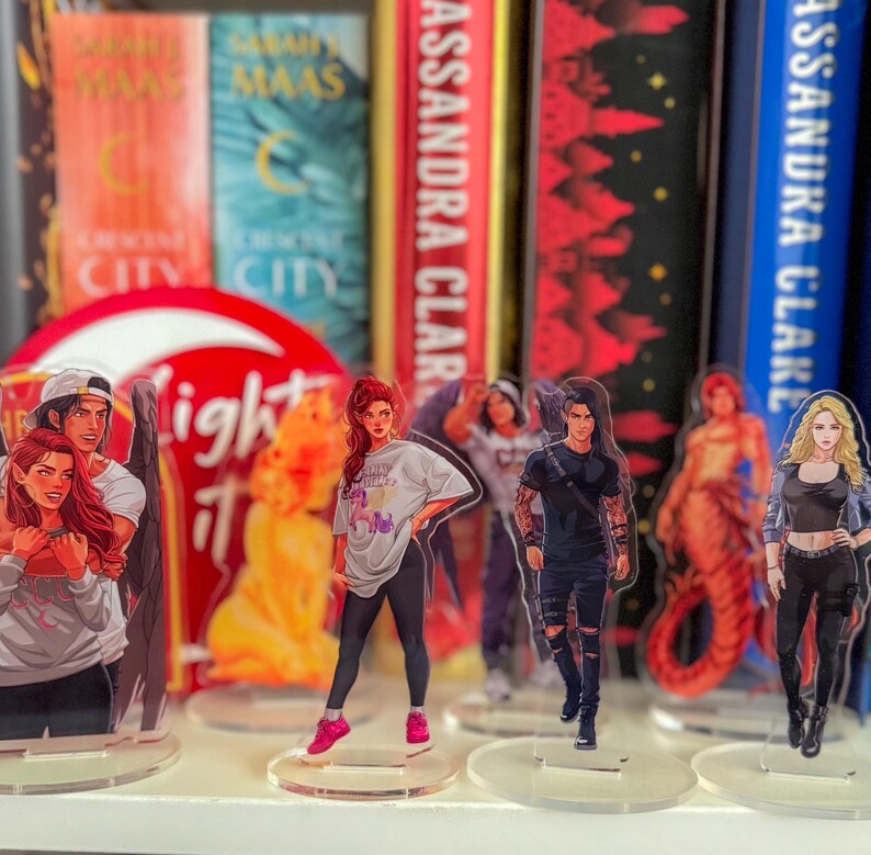 May include: Acrylic character figures of various fantasy characters, standing on clear circular bases. The figures depict characters in casual clothing, with vibrant colors and detailed designs. Bookshelves are in the background.