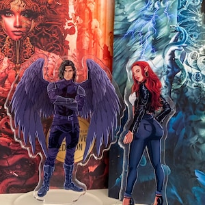 May include: Two acrylic standees featuring fantasy characters. One depicts a male figure with large purple wings, wearing a dark outfit. The other shows a female figure with red hair, a black jacket, and dark jeans. Both stand on clear bases.
