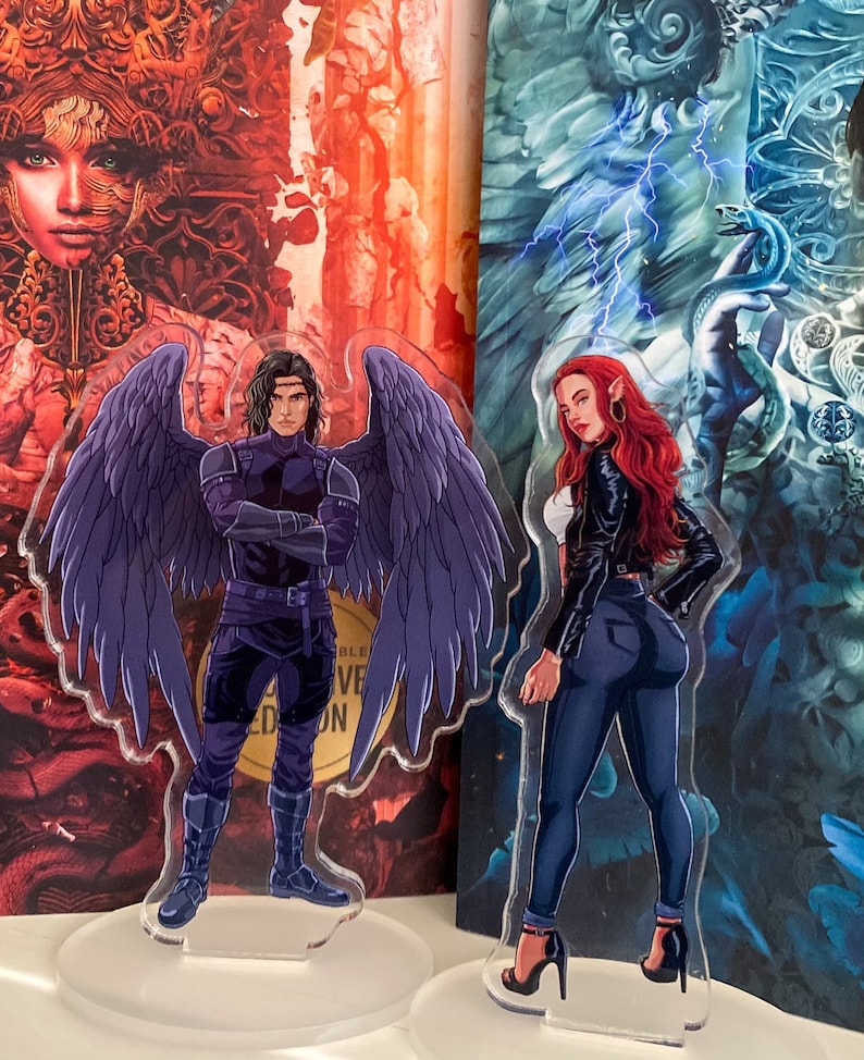 May include: Two acrylic standees featuring fantasy characters. One depicts a male figure with large purple wings, wearing a dark outfit. The other shows a female figure with red hair, a black jacket, and dark jeans. Both stand on clear bases.