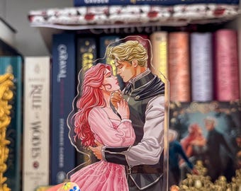 May include: A clear acrylic standee with a romantic illustration of a couple. The woman has long pink hair and wears a pink dress. The man has blond hair and wears a dark outfit. The standee is held in front of a bookshelf filled with books.