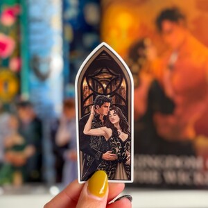 May include: A sticker with a romantic illustration of a couple embracing. The artwork shows a man and woman in formal attire, set against a gothic arch background. The sticker has a white border and is held by a hand with yellow nail polish.