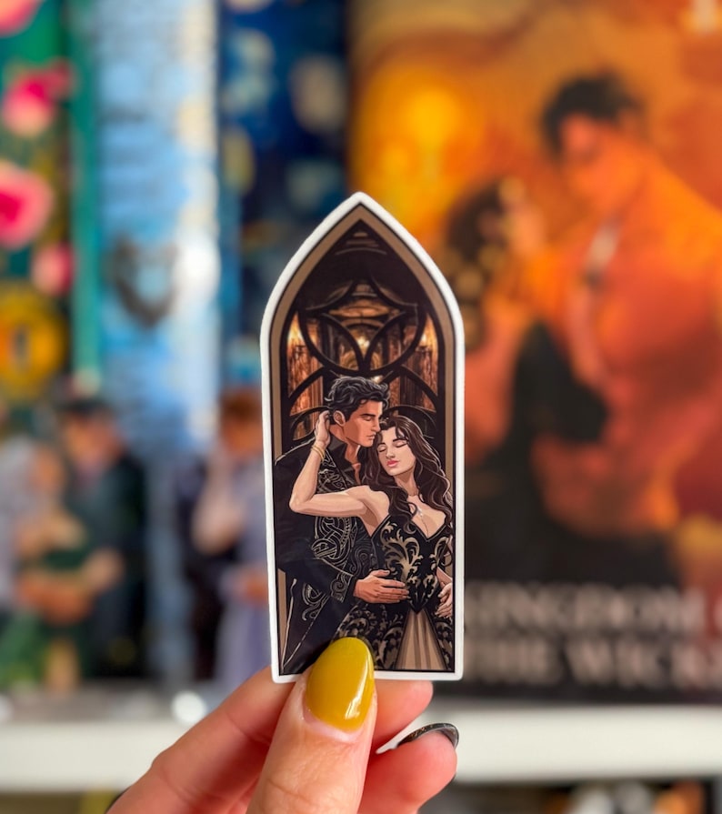 May include: A sticker with a romantic illustration of a couple embracing. The artwork shows a man and woman in formal attire, set against a gothic arch background. The sticker has a white border and is held by a hand with yellow nail polish.