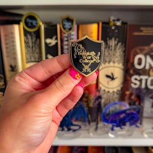 Könnte beinhalten: Ein schwarzer und goldener schildförmiger Anstecker mit dem Text "Basgiath War College" und gekreuzten Schwertern. Der Anstecker wird vor einem Bücherregal mit Büchern und dekorativen Lesezeichen gehalten.