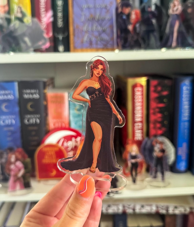 May include: A clear acrylic standee featuring a woman in a black gown with a high slit and red hair. The standee is held in front of a bookshelf filled with books and other figurines. The woman is wearing a strapless dress.