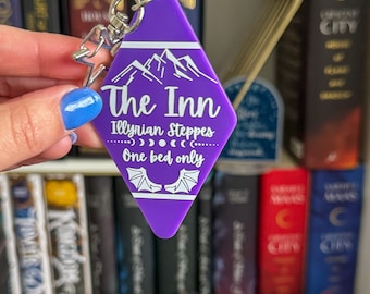 May include: A purple diamond-shaped keychain with the text "The Inn" and "Illyrian Steppes" in white. The keychain features a mountain graphic and the phrase "One bed only". A silver key ring is attached.
