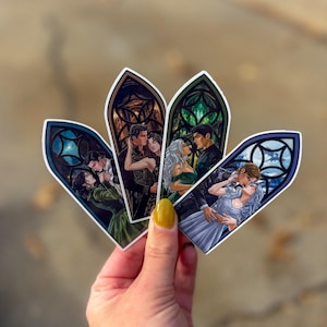 May include: Four stickers with romantic illustrations in stained-glass window frames. Each sticker shows a couple embracing, with different colour schemes and designs. The stickers are held in a hand with yellow nail varnish.