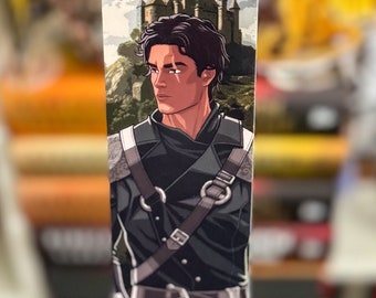 May include: A bookmark featuring a male character with dark hair and a black outfit, set against a backdrop of a castle and a blue dragon. The bookmark is held in a hand with blue nail polish.