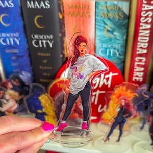 May include: Clear acrylic standee featuring a stylized illustration of a woman with red hair, wearing a white t-shirt with the text "FLLY BLUFF" and black pants. She is wearing pink sneakers. The standee is in front of a bookshelf with books.