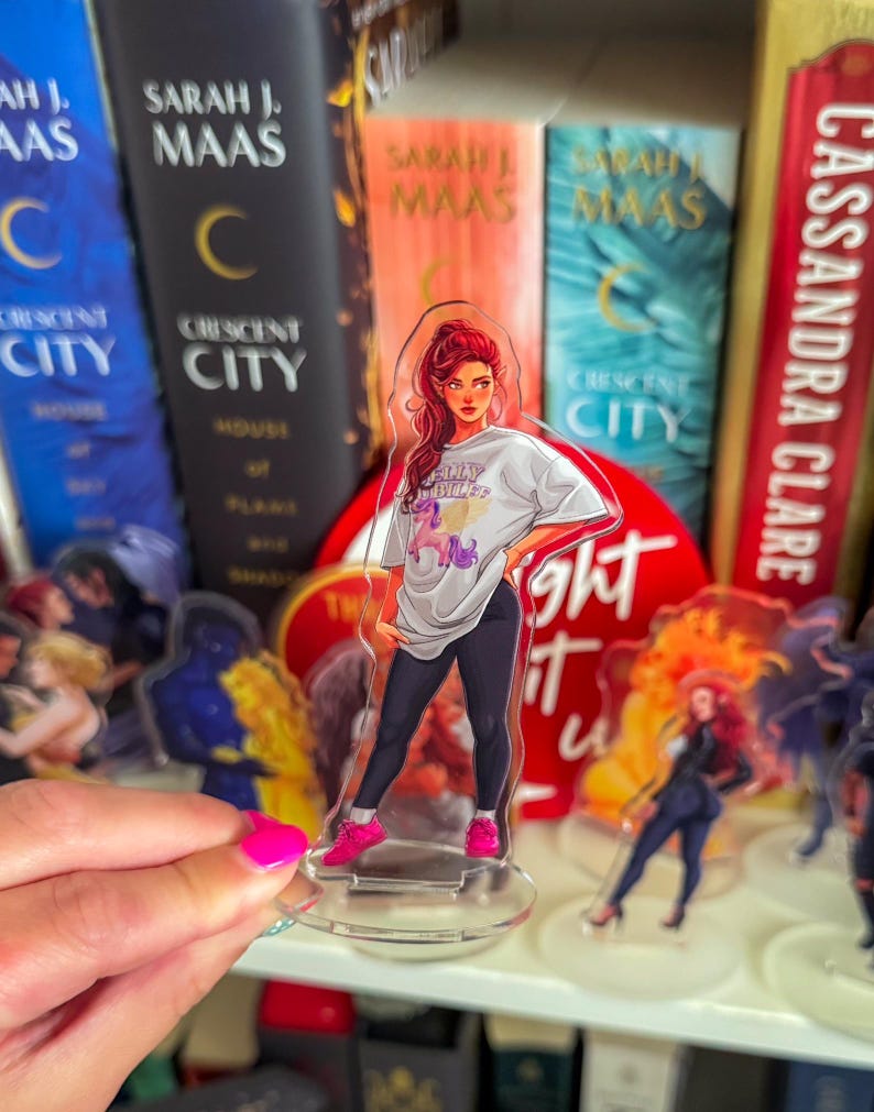 May include: Clear acrylic standee featuring a stylized illustration of a woman with red hair, wearing a white t-shirt with the text "FLLY BLUFF" and black pants. She is wearing pink sneakers. The standee is in front of a bookshelf with books.