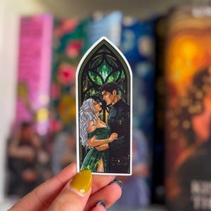 May include: A sticker with a romantic illustration of a couple in a stained-glass window design. The woman has long, silver hair and wears a green dress. The man is in a black suit. The sticker is held by a hand with yellow nail polish.