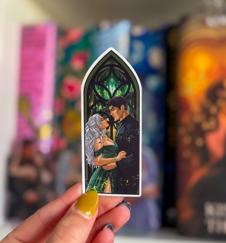 May include: A sticker with a romantic illustration of a couple in a stained-glass window design. The woman has long, silver hair and wears a green dress. The man is in a black suit. The sticker is held by a hand with yellow nail polish.