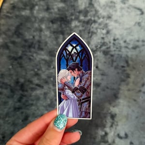 May include: A die-cut sticker featuring an arched window frame design. Inside the frame, a person with blonde hair in a white dress embraces a person in dark metal armour. The background shows a dark blue night sky with a moon and silhouettes.