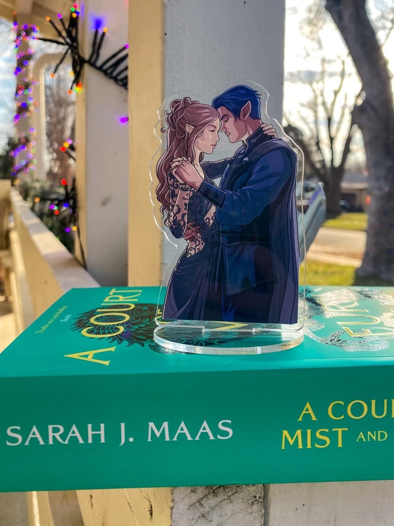 K&ouml;nnte beinhalten: Eine transparente Acrylfigur eines tanzenden Paares mit blauen und braunen Haaren auf einem transparenten Sockel. Die Figur steht auf einem t&uuml;rkisfarbenen Buch mit dem Titel "A Court of Mist and Fury" von Sarah J. Maas.