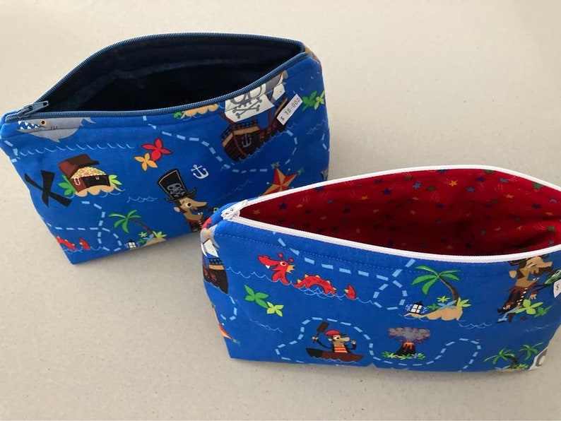 May include: Two blue zippered pouches with a pirate map design. The pouches feature a treasure map pattern with pirate ships, treasure chests, and palm trees. One pouch has a navy blue interior, and the other has a red interior.