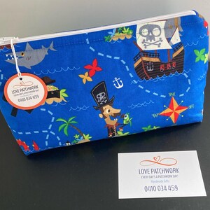 May include: Blue fabric zippered pouch with a pirate map design. The pouch features illustrations of a pirate ship, a shark, a pirate, and a treasure island. The text "LOVE PATCHWORK" and a phone number are visible on a tag attached to the zipper.