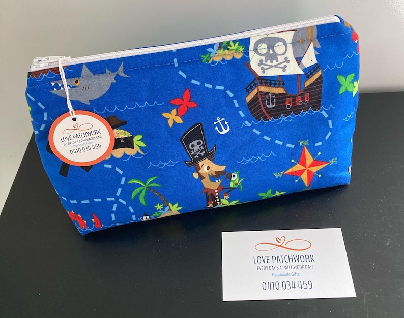 May include: Blue fabric zippered pouch with a pirate map design. The pouch features illustrations of a pirate ship, a shark, a pirate, and a treasure island. The text "LOVE PATCHWORK" and a phone number are visible on a tag attached to the zipper.