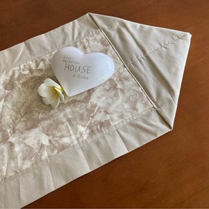 May include: Beige table runner with a leaf pattern in the center and a solid beige border. A white heart-shaped ornament with the text "Love Makes Our HOUSE A Home" and a white flower are on the runner.