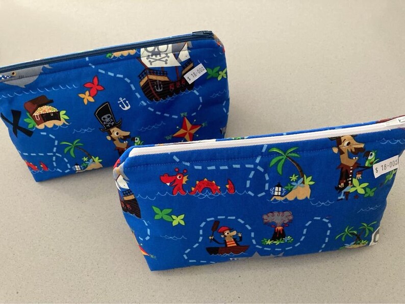 May include: Two blue zippered pouches with a pirate map design. The fabric features a treasure map with cartoon pirates, ships, treasure chests, and palm trees. One pouch has a blue zipper, and the other has a white zipper.