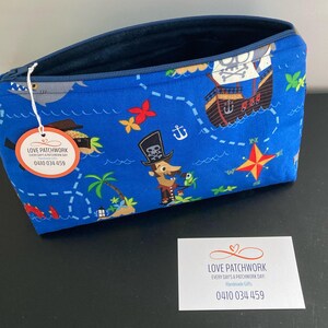 May include: A blue zippered pouch with a pirate map design. The fabric features a pirate ship, treasure chest, and cartoon pirate characters. A tag with the text "LOVE PATCHWORK" and a phone number is attached.