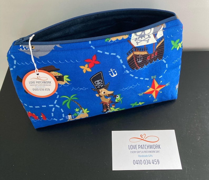 May include: A blue zippered pouch with a pirate map design. The fabric features a pirate ship, treasure chest, and cartoon pirate characters. A tag with the text "LOVE PATCHWORK" and a phone number is attached.