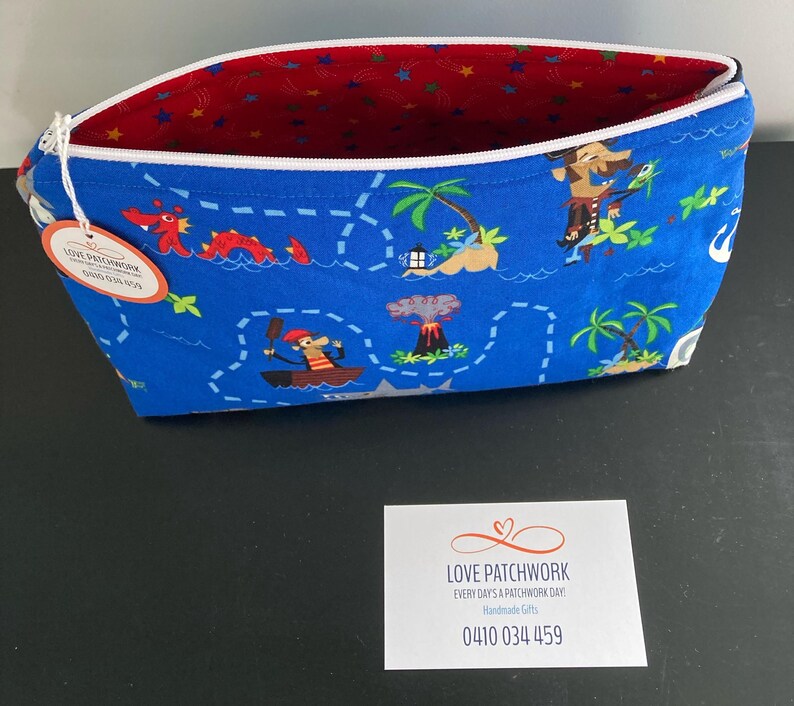 May include: A blue fabric zippered pouch with a pirate map design. The pouch features illustrations of pirates, islands, and sea creatures. The interior is red with a star pattern. A tag reads "LOVE PATCHWORK".