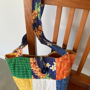 May include: A patchwork tote bag with a blue, green, yellow, orange, and white floral print. The bag has a single button closure and a long strap.