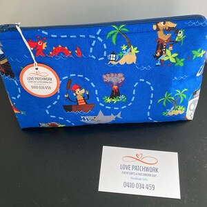 May include: Blue zippered pouch with a pirate map design featuring cartoon pirates, palm trees, and a shark. The pouch has a tag that says "LOVE PATCHWORK" and a phone number.