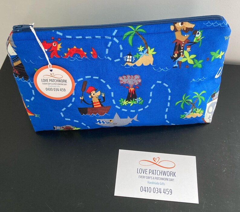 May include: Blue zippered pouch with a pirate map design featuring cartoon pirates, palm trees, and a shark. The pouch has a tag that says "LOVE PATCHWORK" and a phone number.