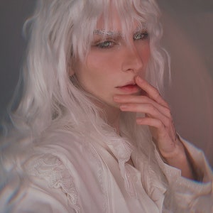 May include: A person with long, wavy white hair and pale skin. They are wearing a white, ruffled shirt with lace detailing. The person is touching their lips with their fingers. The background is a soft, blurred pink.