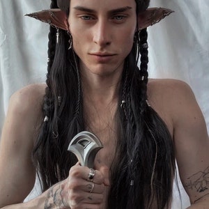 Pode incluir: Uma pessoa com cabelo comprido e preto, tranças e orelhas pontudas, segurando uma espada de prata. A pessoa tem tatuagens nos braços e dedos, e usa anéis. O fundo é um lençol branco.