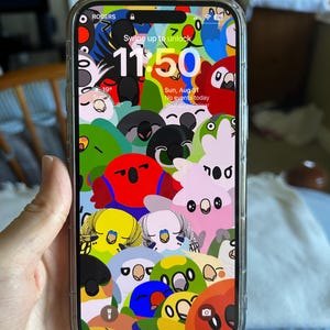 Puede incluir: Un smartphone con una funda transparente muestra un fondo de pantalla de loros de dibujos animados. La hora es 11:50, con la fecha y la temperatura mostradas. El teléfono se sostiene en una mano.