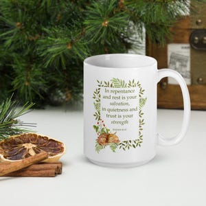 May include: A white ceramic mug featuring a green botanical frame with fern leaves and red berries. Inside the frame is the text: In repentance and rest is your salvation, in quietness and trust is your strength, Isaiah 30:15. A small fawn is illustrated below the text.