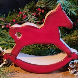 Könnte beinhalten: Ein rot-weißes Holz-Schaukelpferd-Weihnachtsornament. Das Pferd hat einen herzförmigen Ausschnitt und ist von Tannennadeln, roten Beeren und Tannenzapfen umgeben.