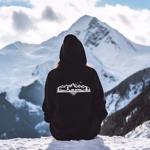 Könnte beinhalten: Ein schwarzer Kapuzenpullover mit einem weißen Bergpanorama-Design auf der Rückseite. Das Design zeigt eine Bergkette mit Bäumen, einem Vogel und einem Reh. Die Berge sind in einem Baumstamm verwurzelt.