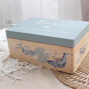 Puede incluir: Caja de recuerdos de madera con tapa azul claro e ilustraciones de ballenas. La caja presenta el nombre "Erea" y detalles de nacimiento. Los lados de la caja están decorados con ballenas, nubes y un barco.
