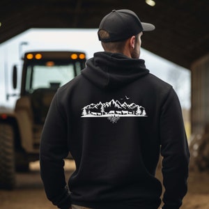 Könnte beinhalten: Schwarzer Hoodie mit einem weißen Aufdruck einer Bergkette, Bäumen und Kühen. Der Aufdruck enthält auch einen Vogel, der über den Bergen fliegt, und eine Blume am Fuße der Berge.