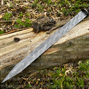 May include: A long, silver sword with a black handle and a patterned blade. The sword is lying on a wooden log.