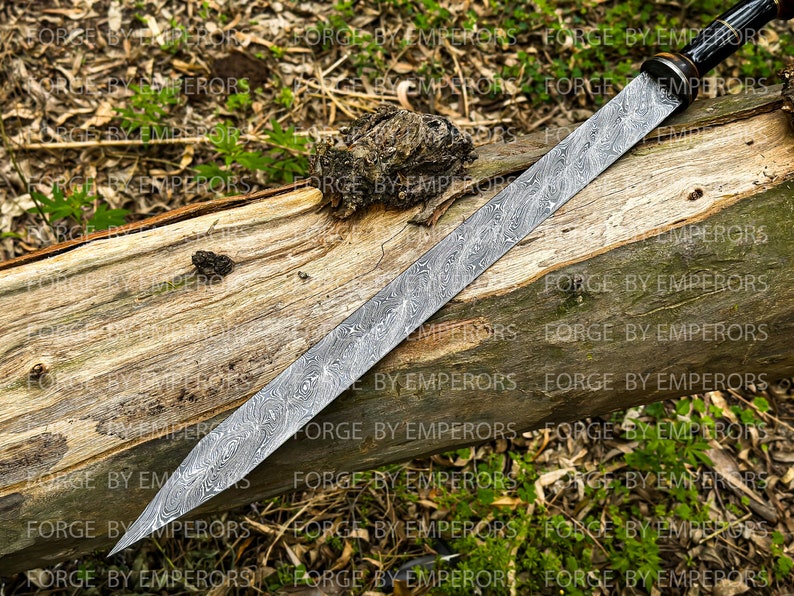 May include: A long, silver sword with a black handle and a patterned blade. The sword is lying on a wooden log.