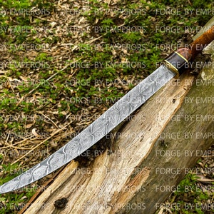 May include: A curved, silver-colored knife with a patterned blade and a brown wooden handle with a gold accent. The knife is resting on a piece of weathered wood.