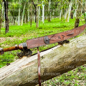 May include: A brown leather scabbard with a wooden-handled sword inside. The scabbard has a decorative design with stitching and a metal accent. The sword is resting on a fallen log in a forest setting.