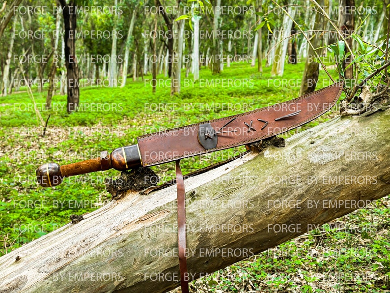 May include: A brown leather scabbard with a wooden-handled sword inside. The scabbard has a decorative design with stitching and a metal accent. The sword is resting on a fallen log in a forest setting.