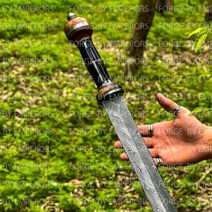 May include: A Damascus steel sword with a wooden handle and a black and silver metal pommel. The sword is held in a person's hand, with the blade pointing towards the camera.