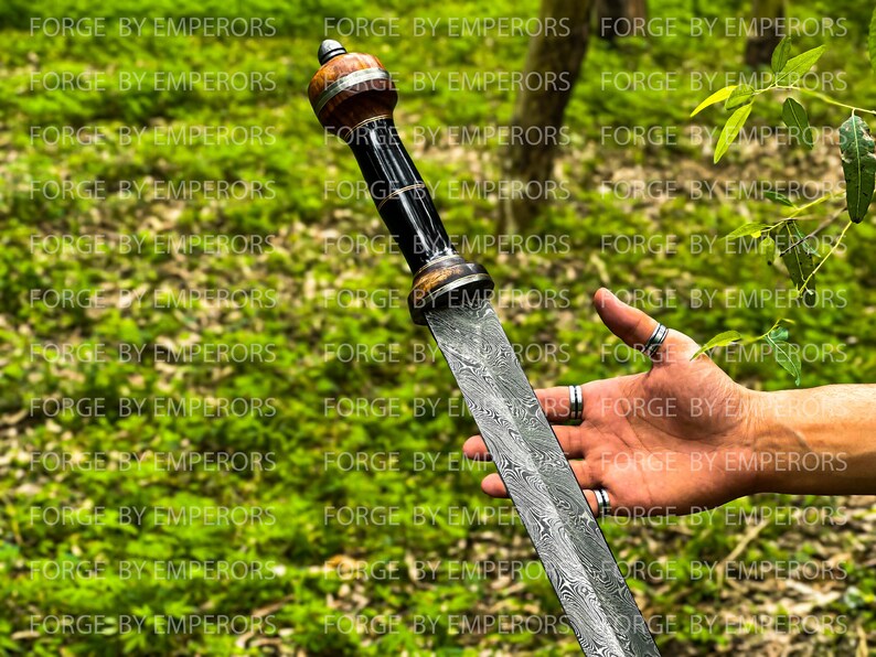 May include: A Damascus steel sword with a wooden handle and a black and silver metal pommel. The sword is held in a person's hand, with the blade pointing towards the camera.