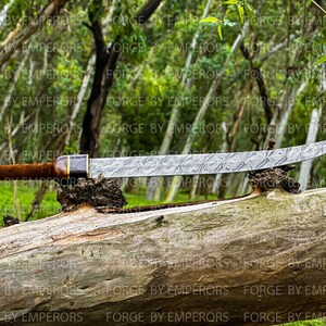 May include: A long, curved, silver-colored sword with a wooden handle and a decorative metal band. The sword is resting on a brown log in a forest setting.