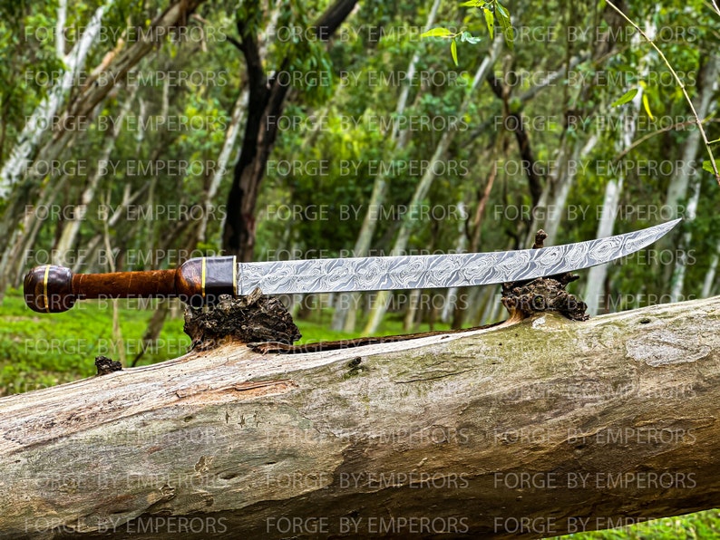 May include: A long, curved, silver-colored sword with a wooden handle and a decorative metal band. The sword is resting on a brown log in a forest setting.