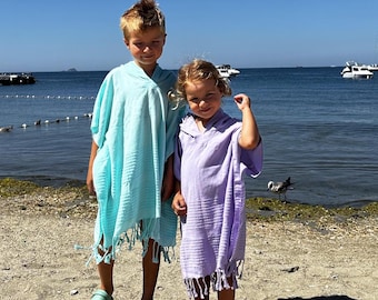 May include: Two children wearing light blue and purple hooded towels stand on a sandy beach with a blue sea in the background.