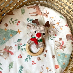 May include: A crocheted reindeer baby rattle with a wooden ring. The reindeer has a red nose, brown antlers, and a red and green scarf. It rests on a white patterned blanket with forest animals and foliage. The blanket is inside a woven basket.