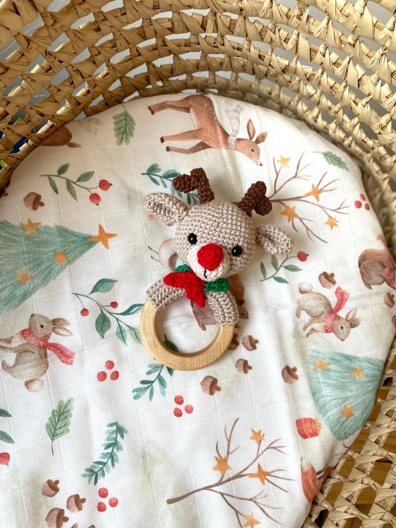 May include: A crocheted reindeer baby rattle with a wooden ring. The reindeer has a red nose, brown antlers, and a red and green scarf. It rests on a white patterned blanket with forest animals and foliage. The blanket is inside a woven basket.