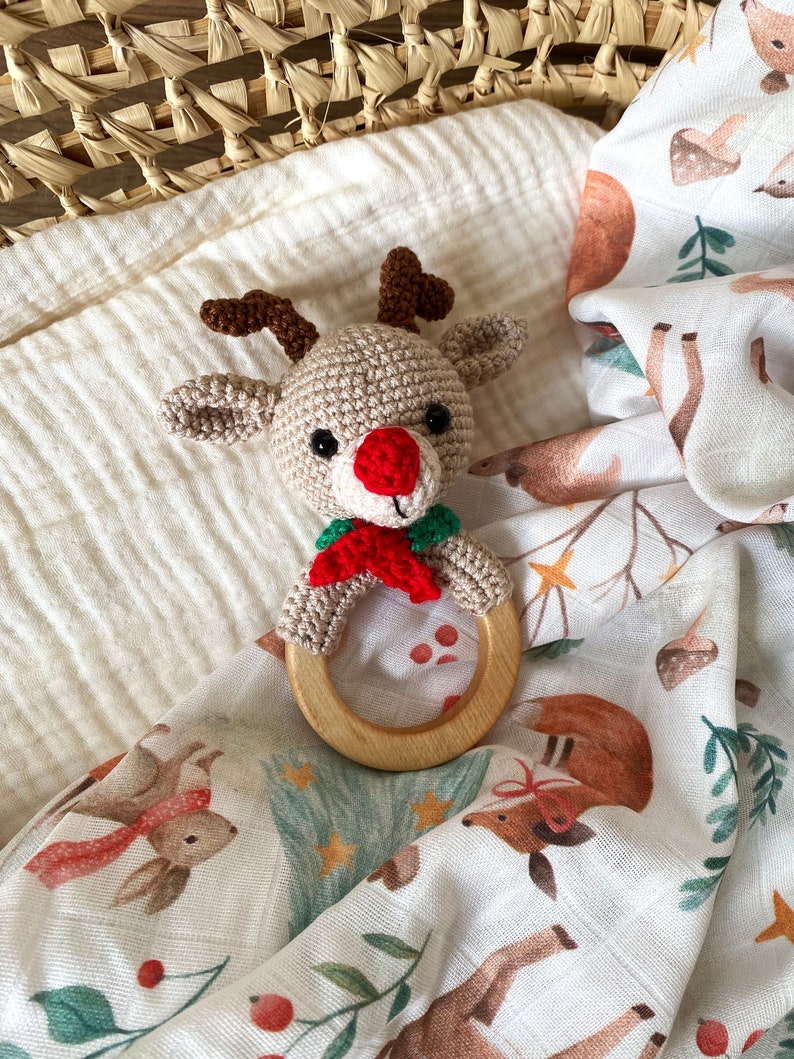 May include: A crocheted reindeer baby rattle with a wooden ring. The reindeer has a beige body, brown antlers, black eyes, and a red nose. It is wearing a green and red scarf. The rattle rests on a white muslin blanket with a woodland animal print.