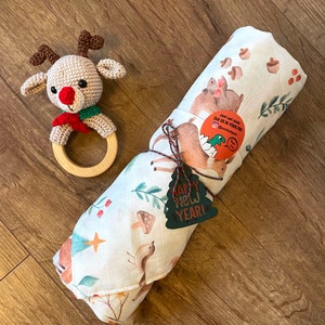 May include: A crocheted reindeer rattle with a wooden ring and a rolled-up white baby blanket featuring woodland animal illustrations. The blanket is tied with twine and a green "Happy New Year!" tag.